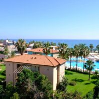 Ein Hotelresort direkt am Mittelmeer mit vielen großen Palmen, einem großen Pool und mehreren kleinen Häusern.