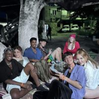 Eine Gruppe von jungen Menschen sitzen bei Nacht, auf Bodenkissen unter einem Baum.