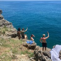 Eine Gruppe von fünf Menschen in Badebekleidung stehen winkend an einer Klippe am Mittelmeer.