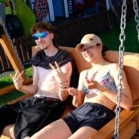 Ein junger Mann mit einer großem blauen Sonnenbrille und ohne T-Shirt sitzt neben einer jungen Frau mit Basecap und Bikini in einer Hollywood-Schauke.